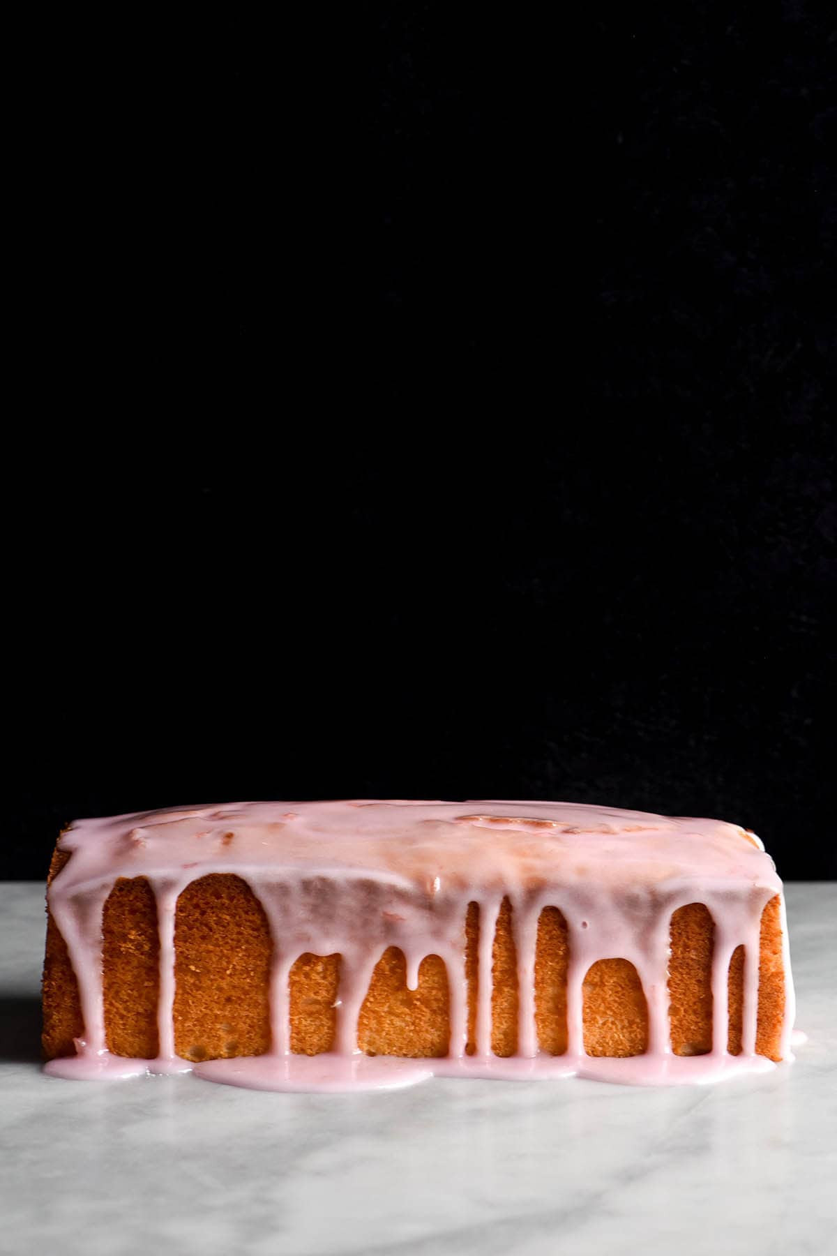A side on view of a loaf of gluten free vanilla cake. It sits on a white marble table with a black background. The loaf is golden brown and drizzled with a light pink icing which snakes down the side of the loaf