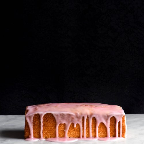 A side on view of a loaf of gluten free vanilla cake. It sits on a white marble table with a black background. The loaf is golden brown and drizzled with a light pink icing which snakes down the side of the loaf