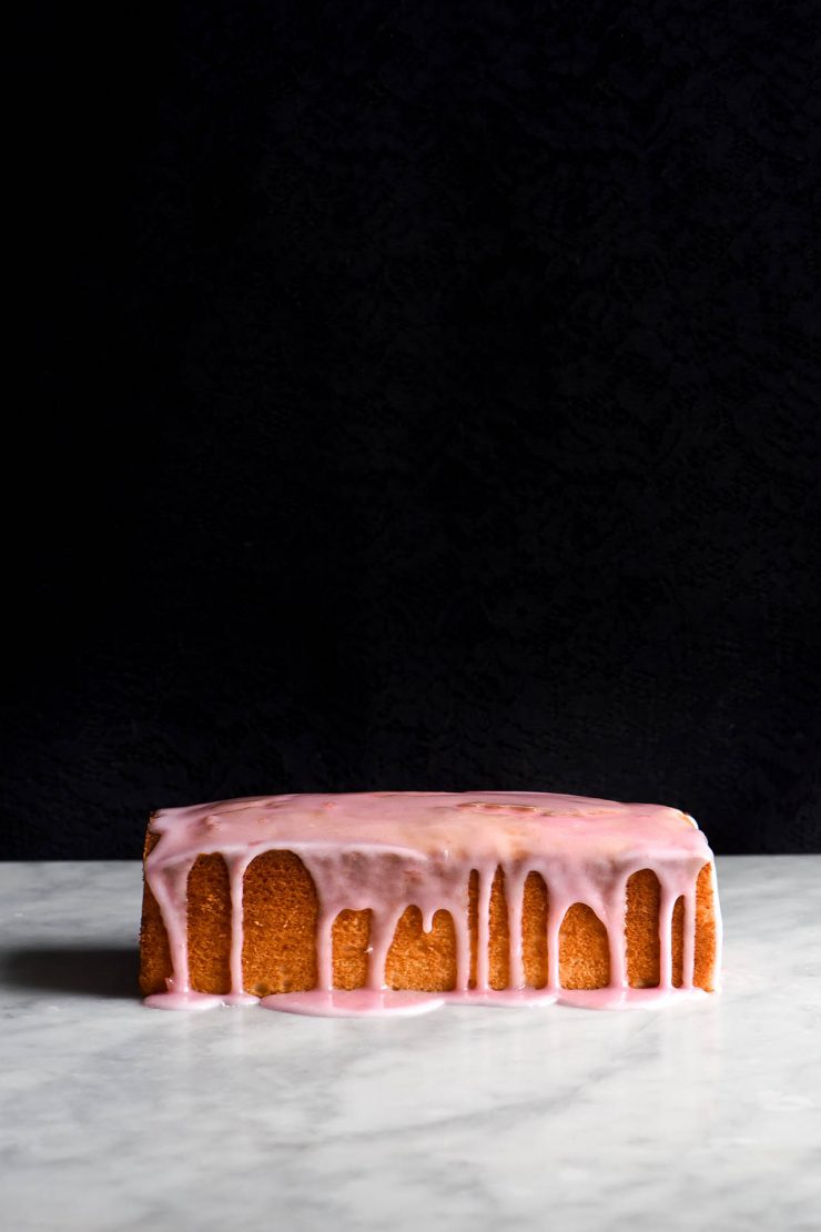 A side on view of a loaf of gluten free vanilla cake. It sits on a white marble table with a black background. The loaf is golden brown and drizzled with a light pink icing which snakes down the side of the loaf