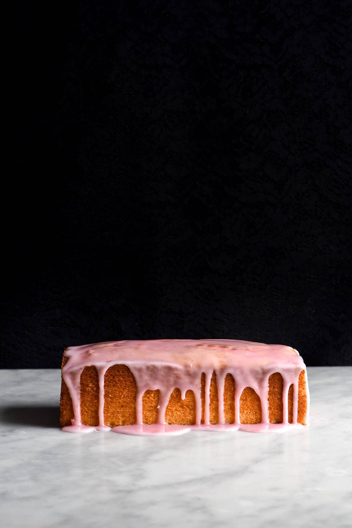 A side on view of a loaf of gluten free vanilla cake. It sits on a white marble table with a black background. The loaf is golden brown and drizzled with a light pink icing which snakes down the side of the loaf