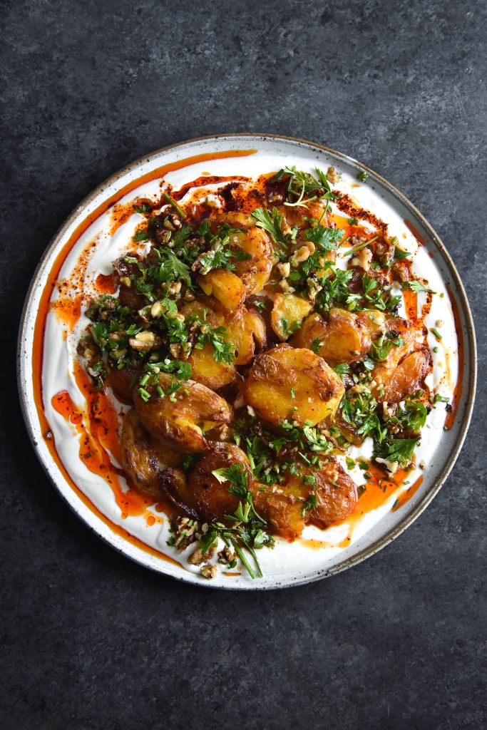 An aerial view of a plate of garlic and chilli oil smothered crispy smashed potatoes on a bed of zingy spiked yoghurt and topped with a walnut herb salsa. The ceramic plate is white and sits on a contrasting dark blue mottled backdrop.