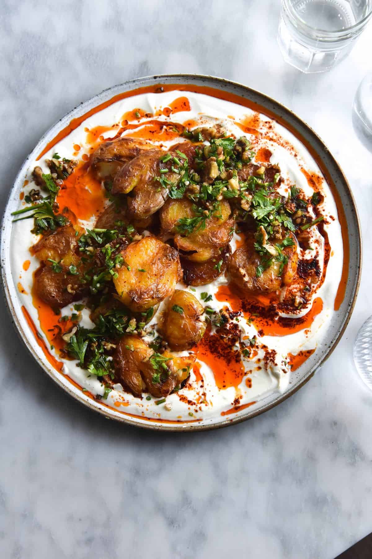 An aerial view of a plate of garlic and chilli oil smothered crispy smashed potatoes on a bed of zingy spiked yoghurt and topped with a walnut herb salsa. The ceramic plate is white, and sits in the centre of the white marble table. Two water glasses sit to the right of the image.