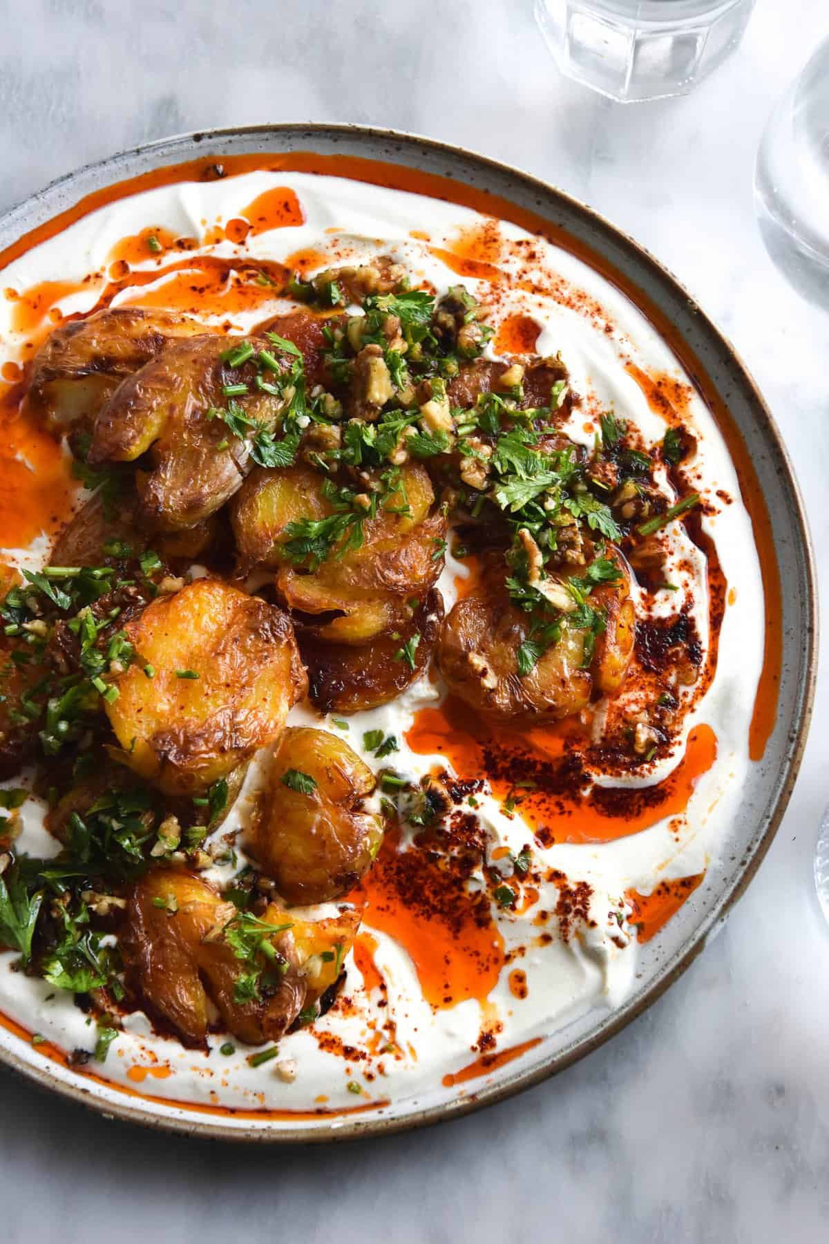 A close up aerial view of a plate of garlic and chilli oil smothered crispy smashed potatoes on a bed of zingy spiked yoghurt and topped with a walnut herb salsa. The ceramic plate is white, and sits in the centre of the white marble table. Two water glasses sit to the right of the image.