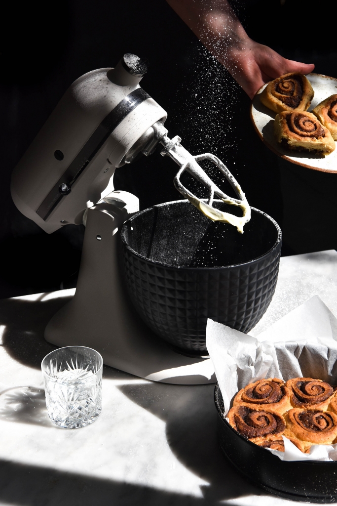 An image of a KitchenAid and gluten free cinnamon scrolls in harsh sunlight. Icing sugar is sprinkled down from the top of the image, and a hand extends out to the right of the image with a plate of cinnamon scrolls