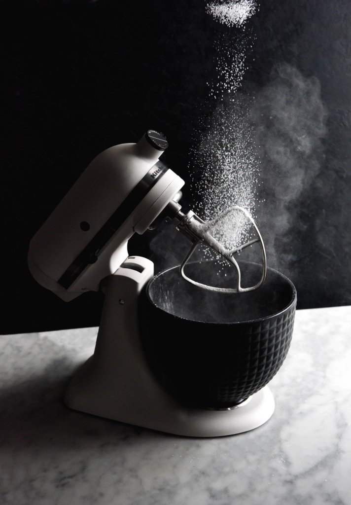 A side on view of a black and white KitchenAid. The KitchenAid sits atop a white marble backdrop against a black backdrop. Flour sprinkles down from the top of the image into the KitchenAid, creating a moody and dramatic shot