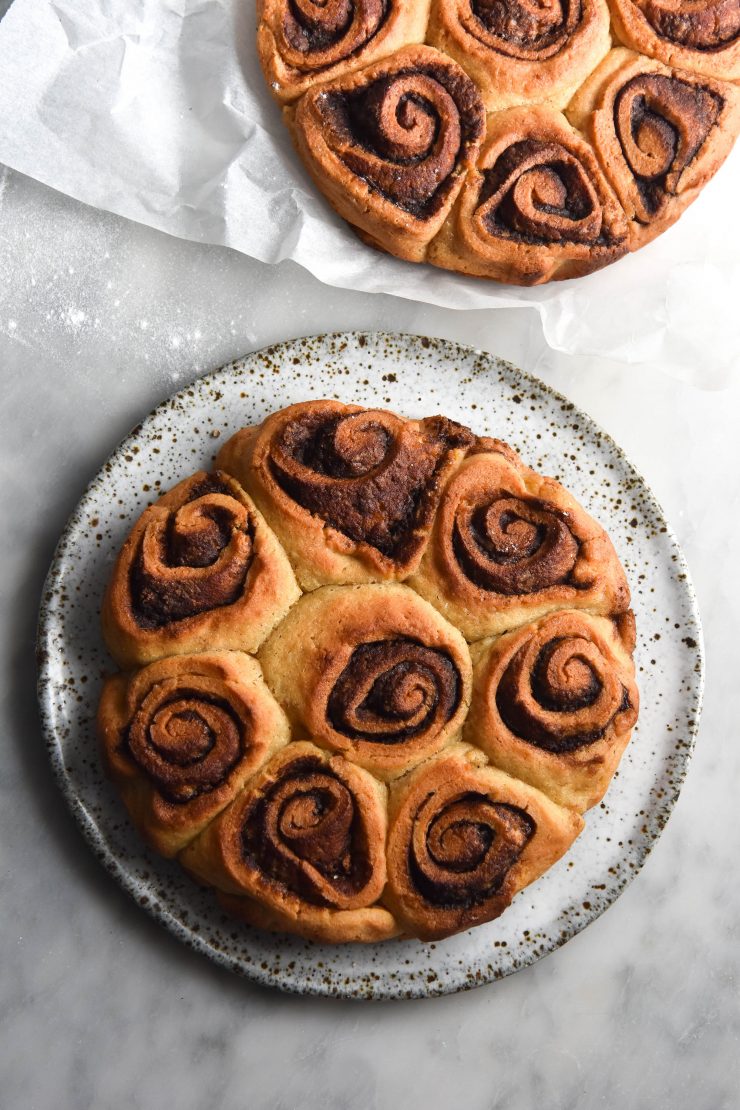 An aerial view of gluten free cinnamon scrolls on a white marble table. One set of scrolls sits atop a white ceramic plate and the other on a sheet of baking paper in the top right hand of the image
