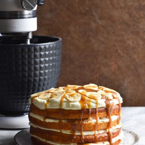 A Gluten free banoffee layer cake sits in the foreground of the image on a ceramic plate against a white marble table. A black and white KitchenAid sits in the background, contrasting against the warm brown mottled tin backdrop