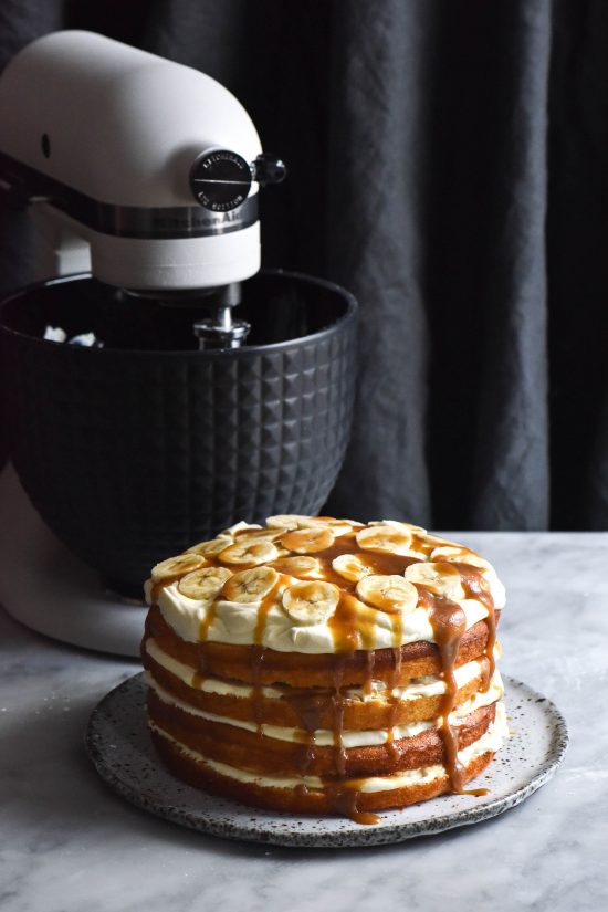 Gluten free banoffee layer cake sits atop a white speckled plate on a white marble backdrop. In the background to the left sits a black and white KitchenAid which contrasts against a black textured linen backdrop