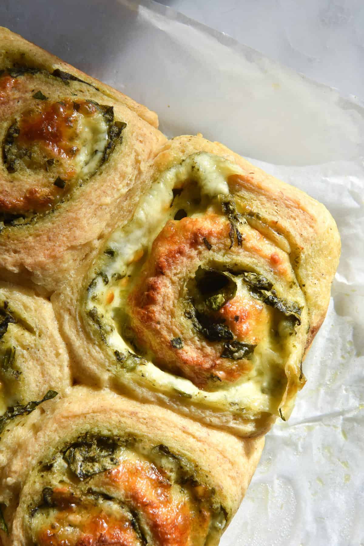 An aerial close up image of freshly baked gluten free pesto scrolls on a white benchtop. The scrolls are golden brown and filled with melty cheese and pesto.