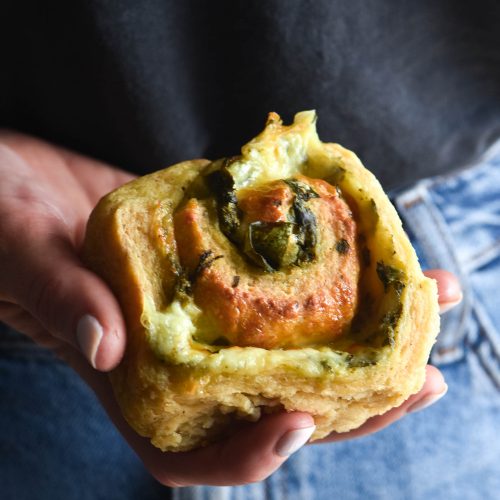 A hand holds out a gluten free pesto and cheese scroll. The person holding the scroll is wearing a denim skirt and a dark top which form the background of the close up image