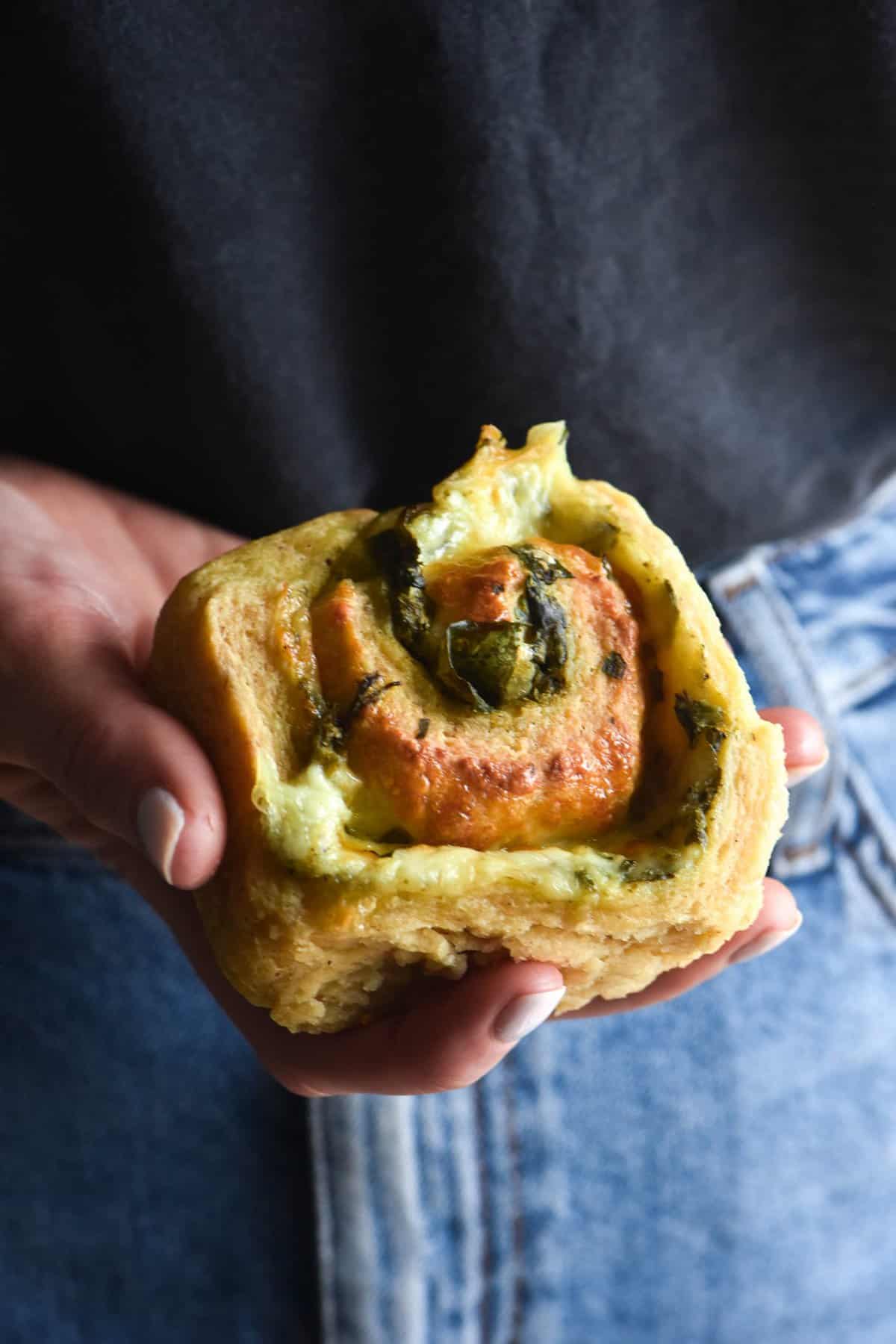 A hand holds out a gluten free pesto and cheese scroll. The person holding the scroll is wearing a denim skirt and a dark top which form the background of the close up image