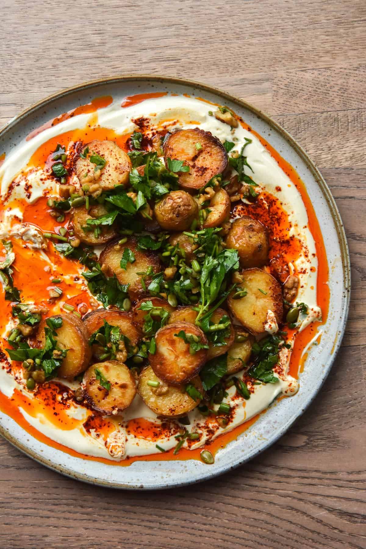 Crispy potatoes with garlic chilli oil, mustard honey yoghurt and a walnut herb salsa sit atop a white ceramic plate against a wooden backdrop