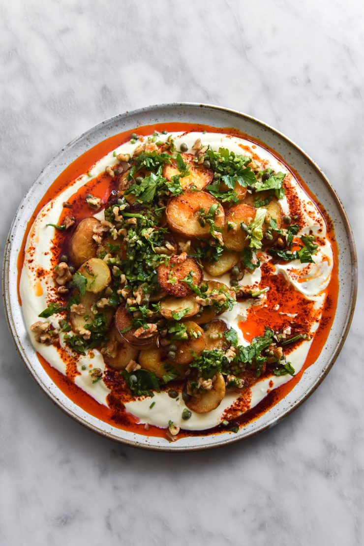 Crispy potatoes with garlic chilli oil, mustard honey yoghurt and a walnut herb salsa on a white ceramic plate atop a white marble table