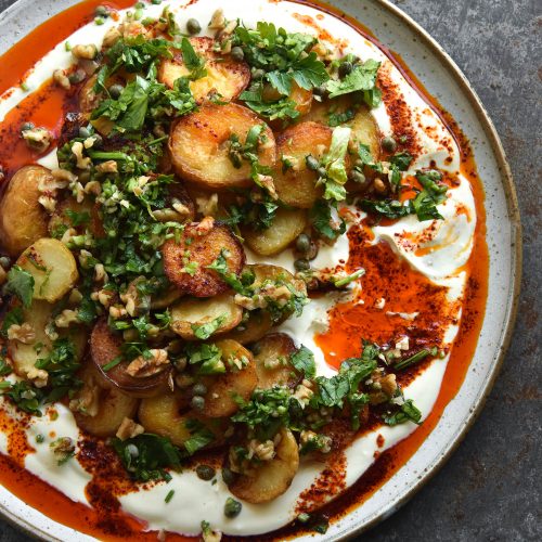 Crispy potatoes with garlic chilli oil, mustard honey yoghurt and a walnut herb salsa sit atop a white ceramic plate on a mottled grey backdrop