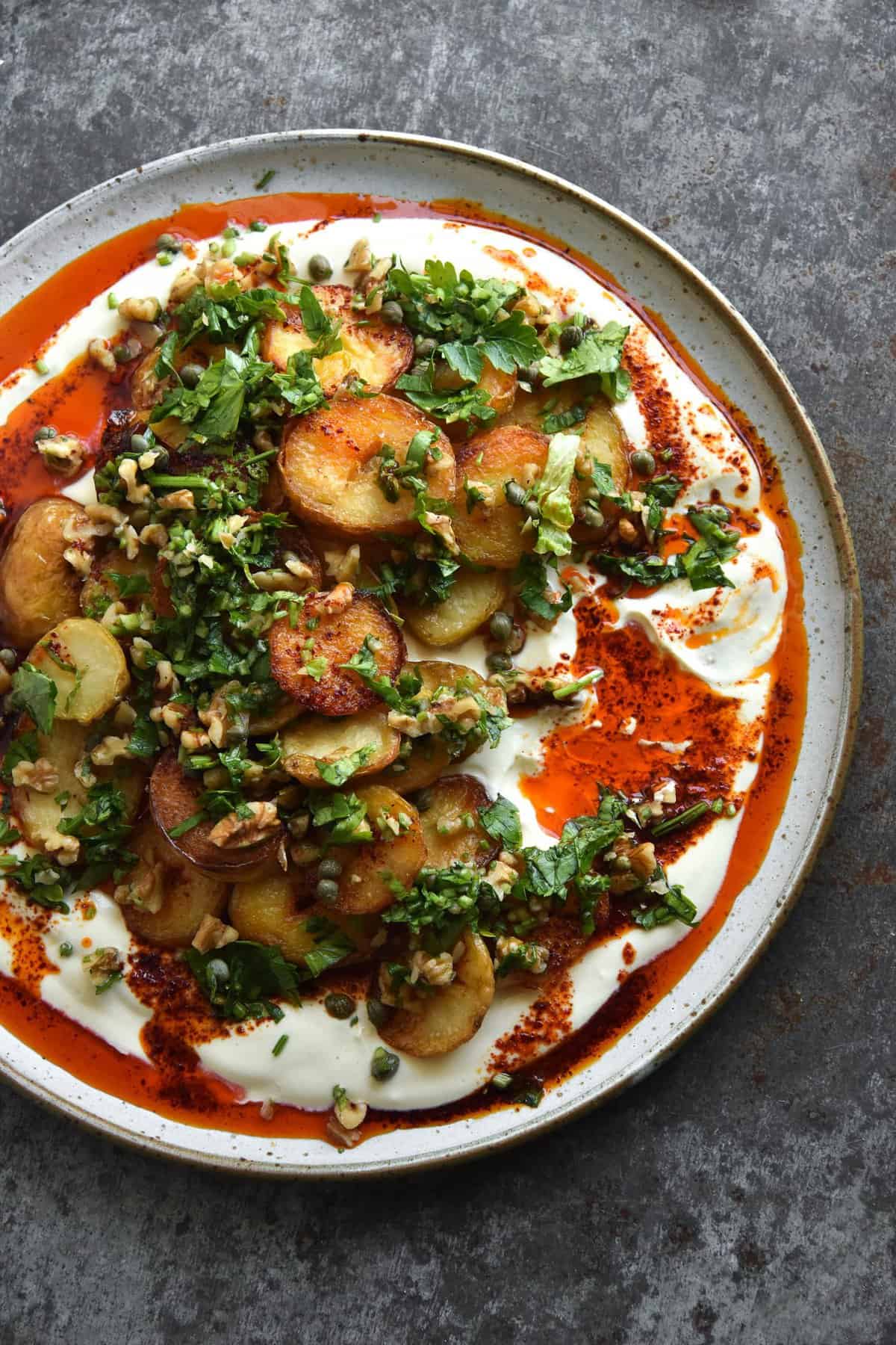 Crispy potatoes with garlic chilli oil, mustard honey yoghurt and a walnut herb salsa sit atop a white ceramic plate on a mottled grey backdrop
