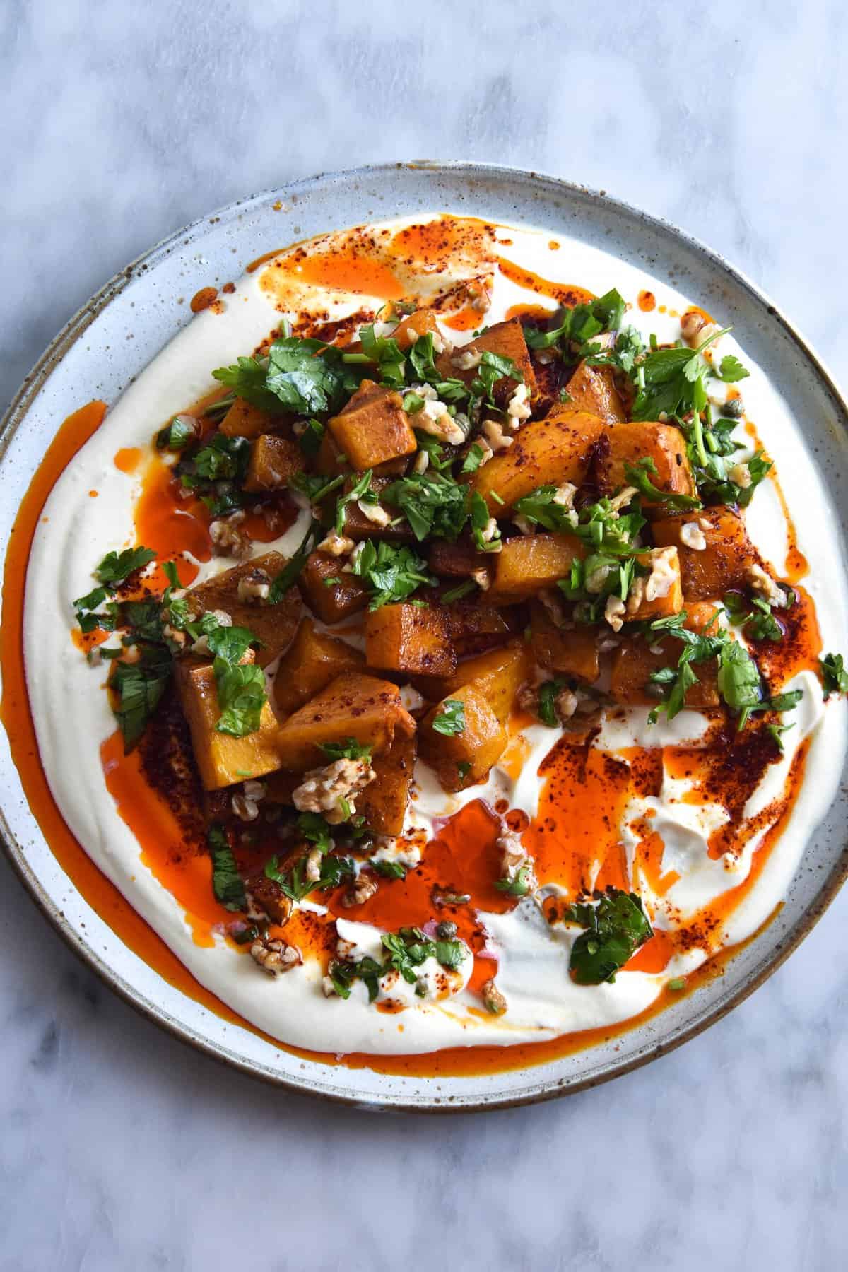 An aerial view of a plate of cinnamon roasted pumpkin atop a honey and mustard yoghurt, drizzled with garlic chilli oil and topped with a herb and nut salsa. The dish sits atop a white ceramic plate on a white marble backdrop