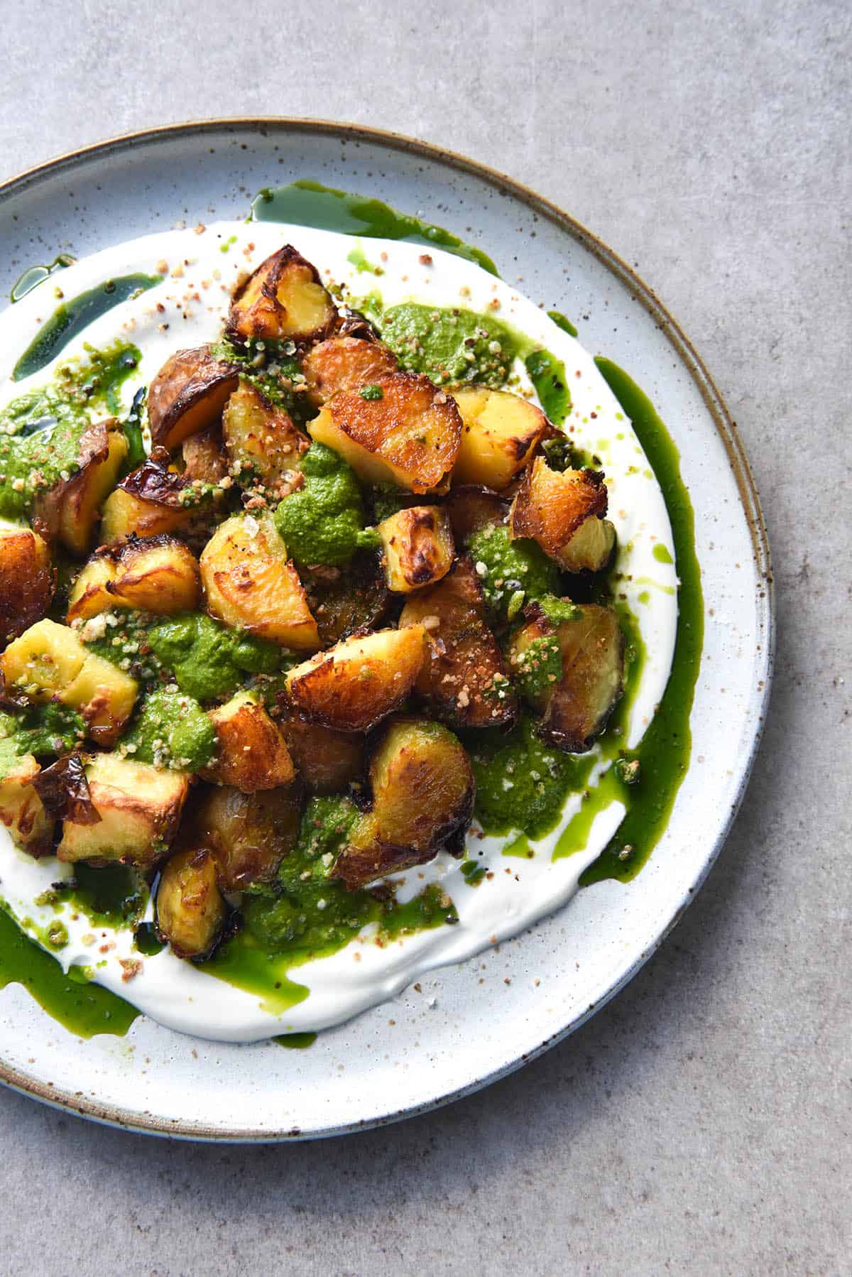 An aerial close up photo of a plate of crispy roast potatoes on a bed of Meredith Goat Cheese dip and topped with FODMAP friendly kale pesto. The green oil from the pesto snakes down the goat cheese spread and onto the plate. The plate is a white ceramic one with a terracotta lip, and it sits atop a light grey stone background