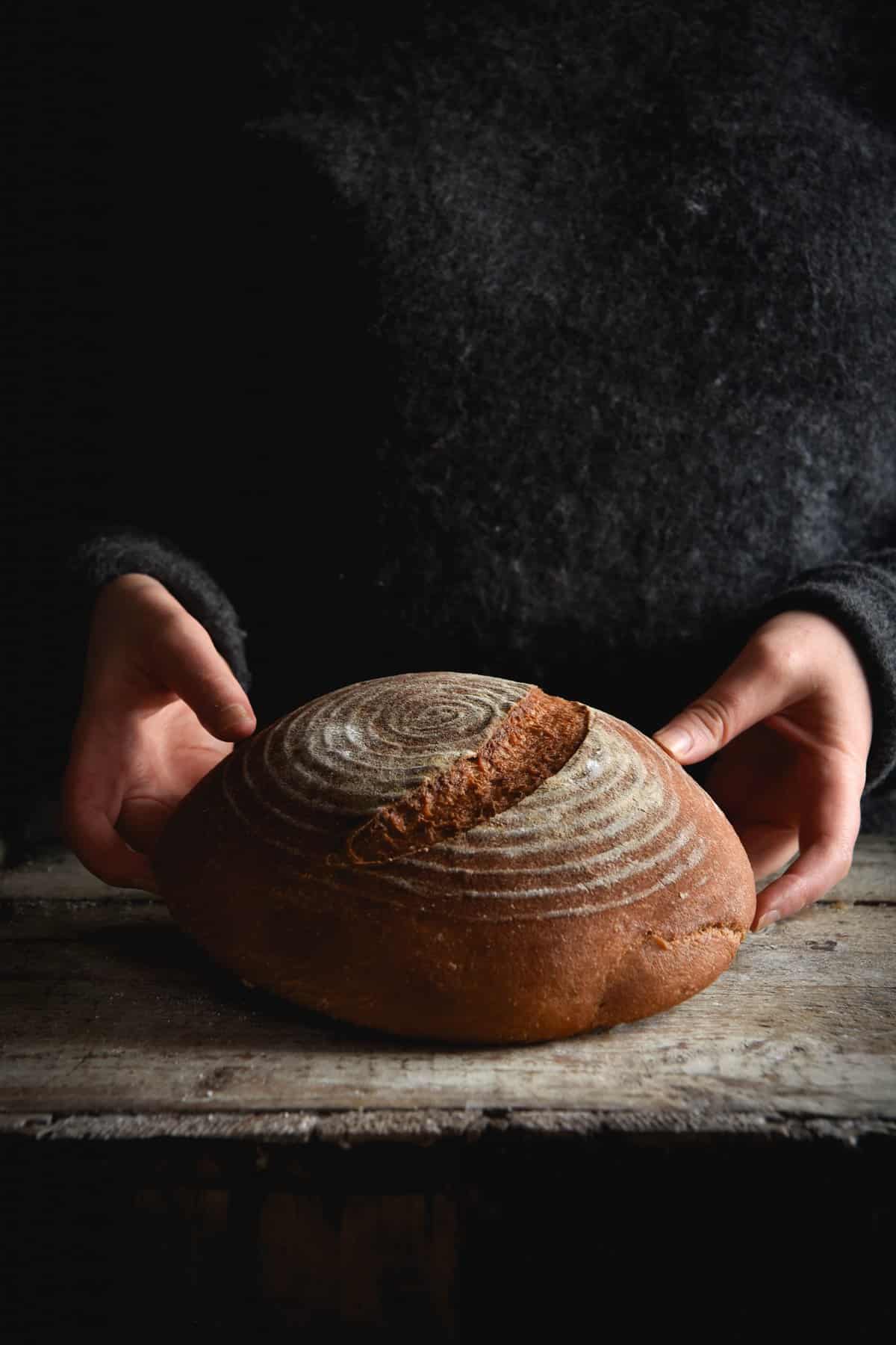 A gluten free sourdough bread boule on a wooden backdrop. A woman in a dark grey jumper stands behind the loaf and holds it up from either side