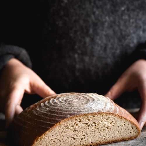 A sliced boule of gluten free sourdough sits on a white wooden backdrop. A person stands behind the loaf wearing a grey woollen jumper, and two hands extends out to hold the loaf on either side.