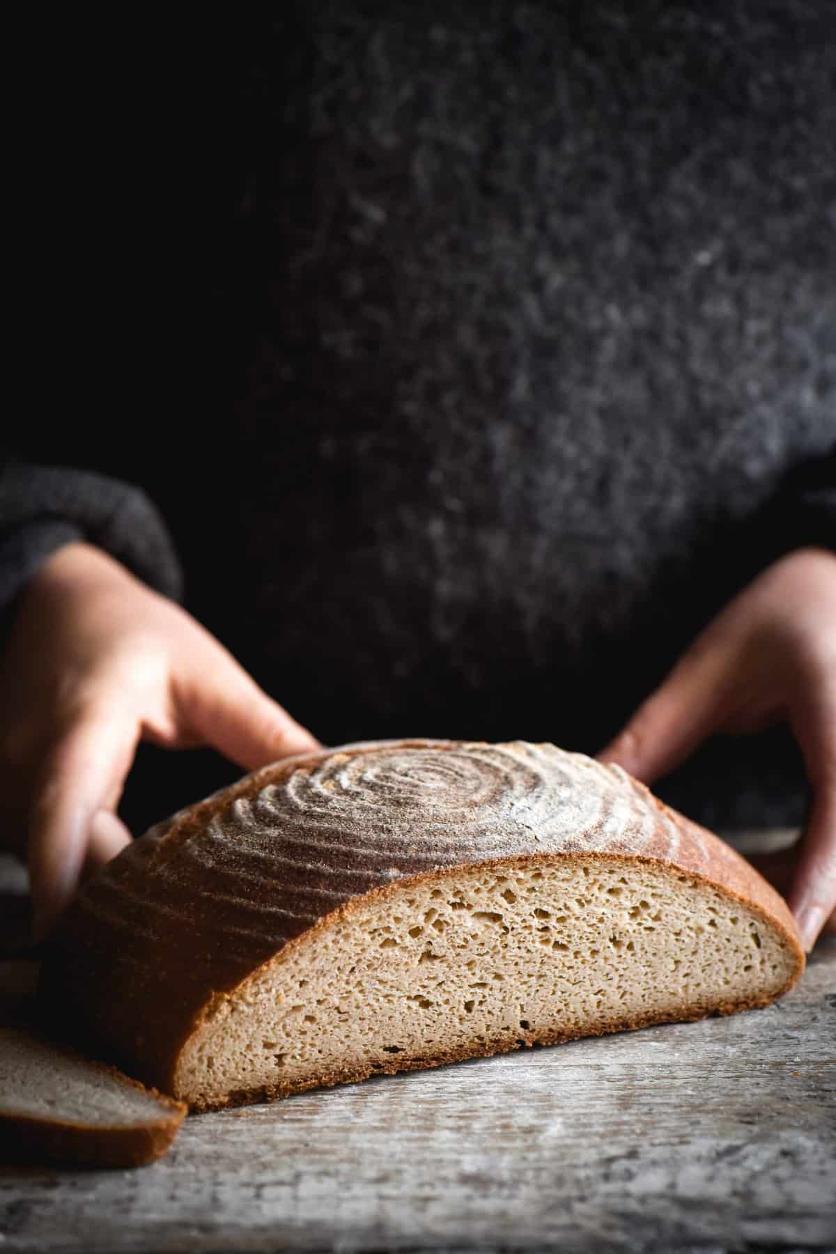 A sliced boule of gluten free sourdough sits on a white wooden backdrop. A person stands behind the loaf wearing a grey woollen jumper, and two hands extends out to hold the loaf on either side.