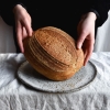 A loaf of gluten free sourdough bread being held up to face the camera. It sits on a speckled white ceramic plate on a white linen tablecloth. The person in the background is wearing a black velvet jacket.