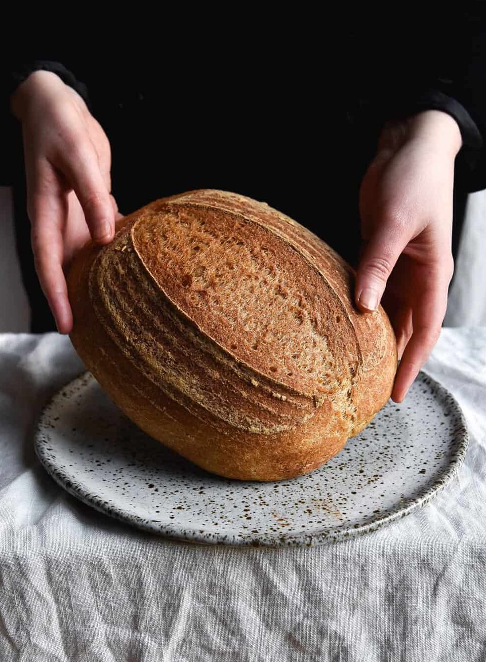 A loaf of gluten free sourdough bread being held up to face the camera. It sits on a speckled white ceramic plate on a white linen tablecloth. The person in the background is wearing a black velvet jacket.