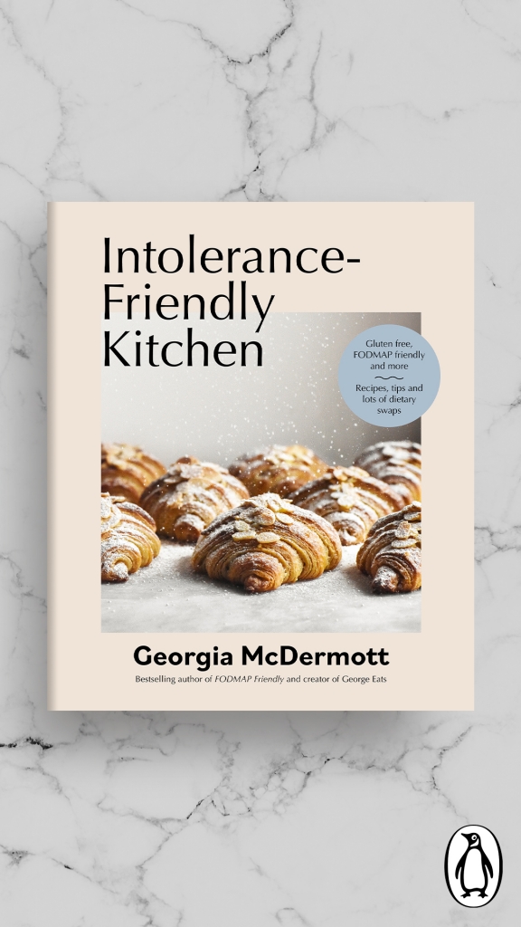 An aerial view of the cookbook cover of Intolerance Friendly Kitchen Cookbook imposed onto a white marble background. The cookbook is a pale pink with a side on shot of gluten free almond croissants being sprinkled with icing sugar on the cover