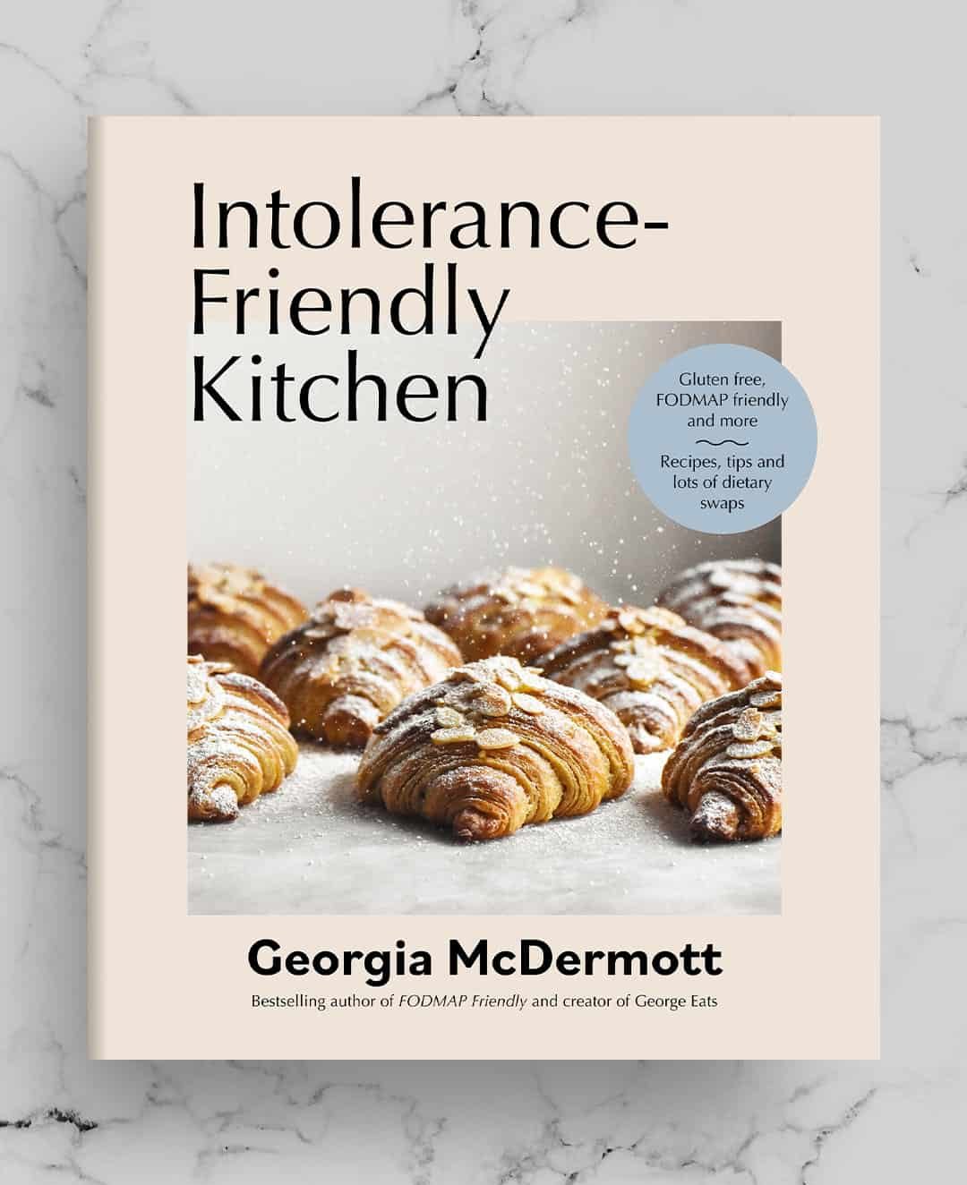 An aerial view of the cookbook cover of Intolerance Friendly Kitchen Cookbook imposed onto a white marble background. The cookbook is a pale pink with a side on shot of gluten free almond croissants being sprinkled with icing sugar on the cover