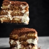 A slice on view of two slices of gluten free, FODMAP friendly tiramisu. One piece of tiramisu is suspended in the air, while the other sits atop a white marble table against a black backdrop