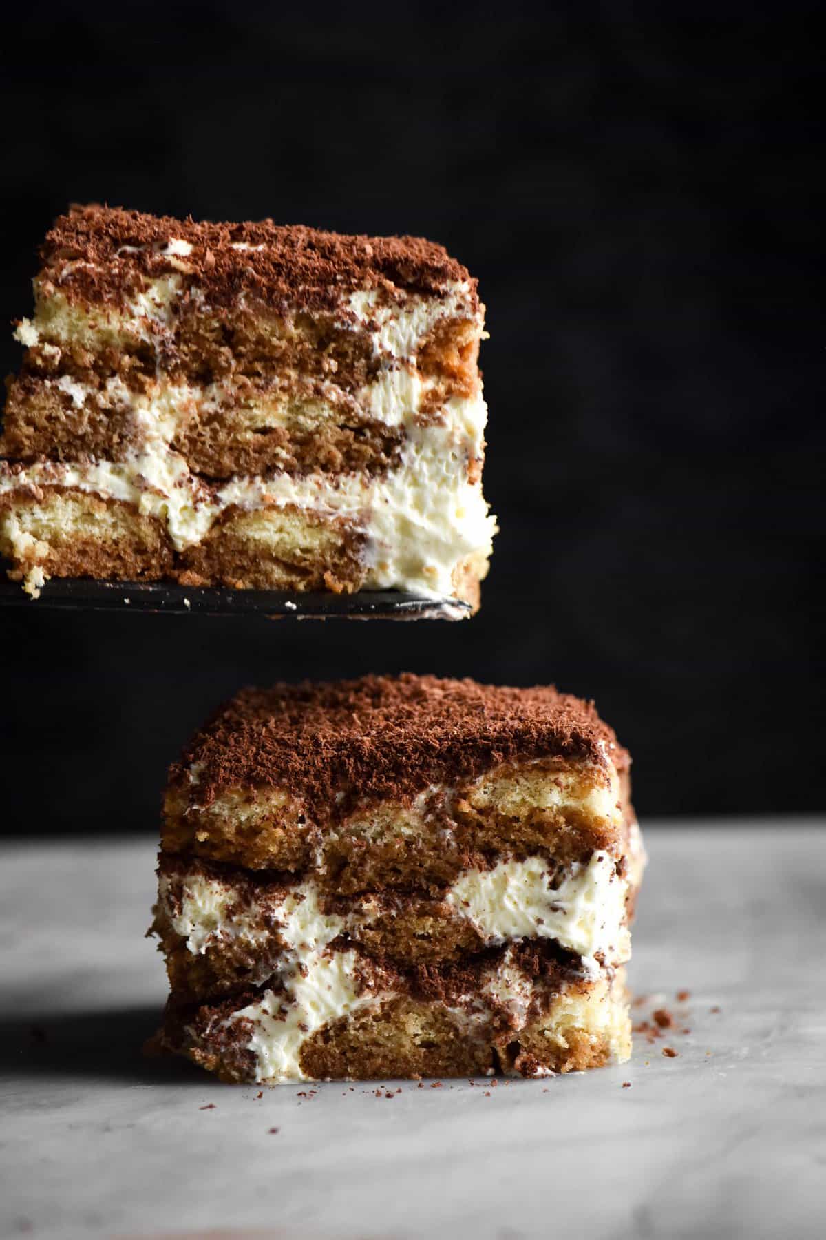 A slice on view of two slices of gluten free, FODMAP friendly tiramisu. One piece of tiramisu is suspended in the air, while the other sits atop a white marble table against a black backdrop