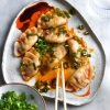 A plate of vegan, gluten free and FODMAP friendly dumplings sit atop a white marble table. The dumplings are pleated, and drizzled with chilli oil, tamari and spring onion greens. Chopsticks rest on the edge of the plate and a small ceramic dish of spring onion greens sits to the left of the dumplings