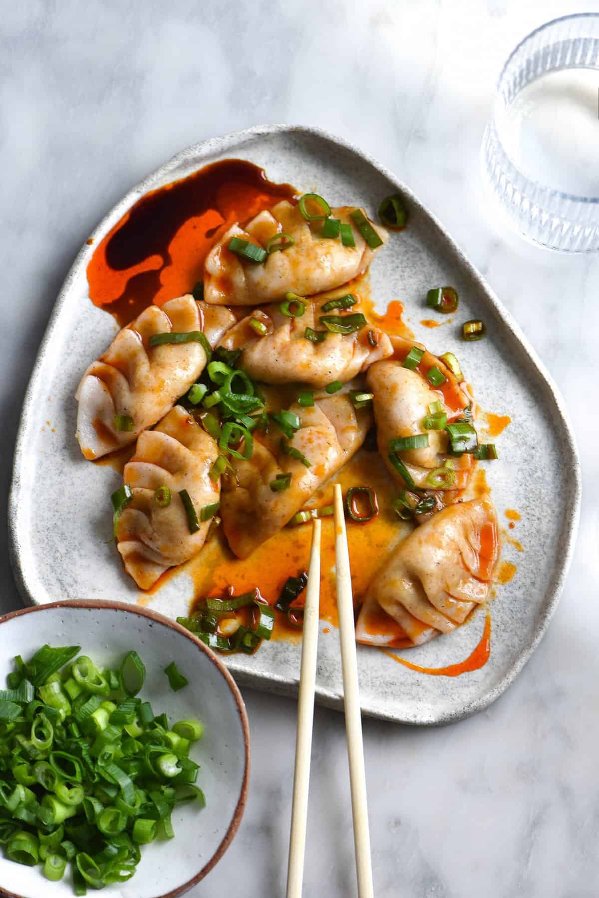 A plate of vegan, gluten free and FODMAP friendly dumplings sit atop a white marble table. The dumplings are pleated, and drizzled with chilli oil, tamari and spring onion greens. Chopsticks rest on the edge of the plate and a small ceramic dish of spring onion greens sits to the left of the dumplings