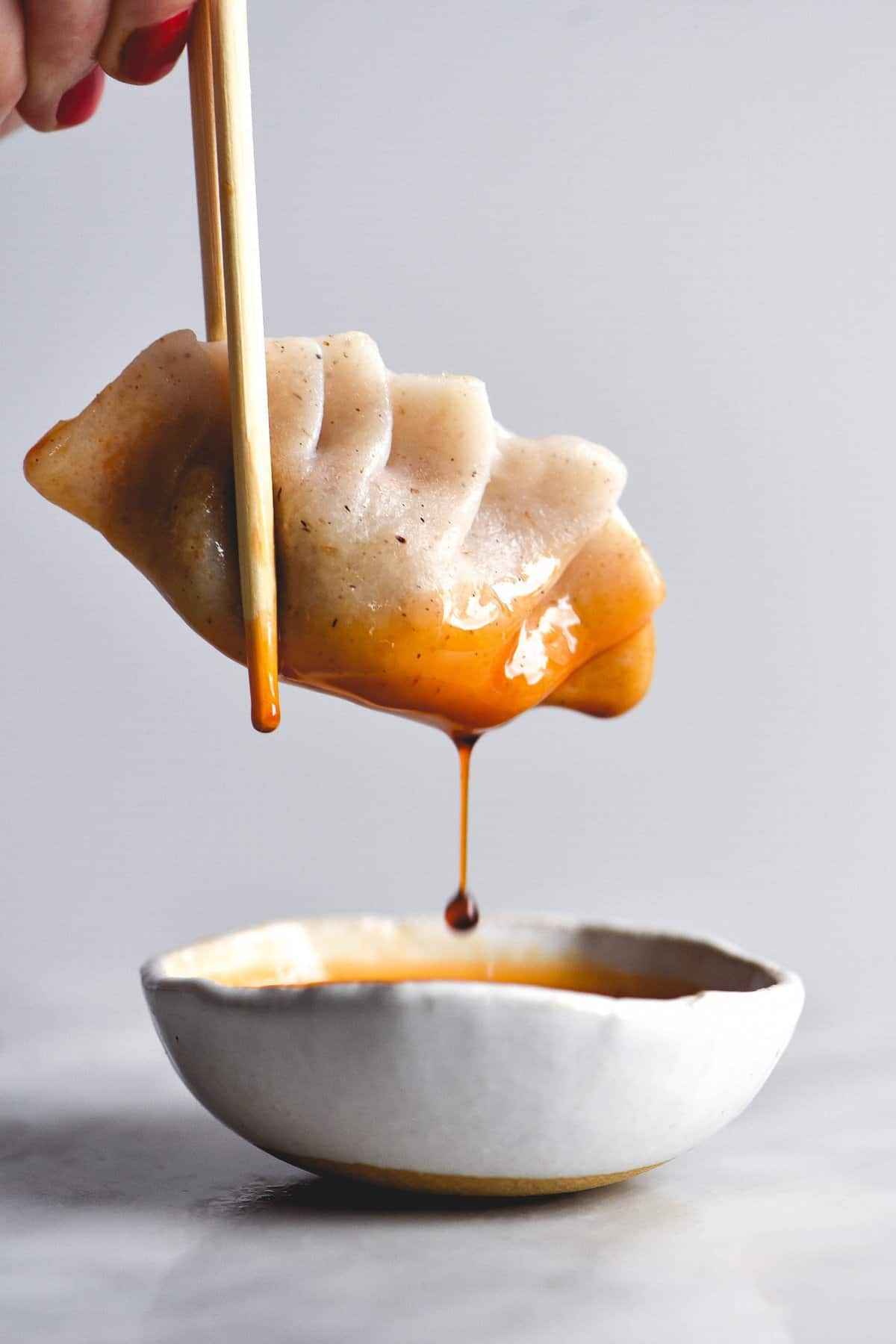 A pleated gluten free dumpling is suspended against a white backdrop by a chopsticks held by a hand to the top left of the image. The dumpling has been dipped into a white ceramic bowl of chilli oil, and a drip of oil is in motion