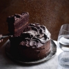 A gluten free chocolate cake sits atop a white marble table against a rusty brown backdrop. A slice of the cake has been removed and is held up by a hand from the left of the image. The whole cake is being sprinkled with snow-like icing sugar, and soft light filters through two water glasses to the right of the image