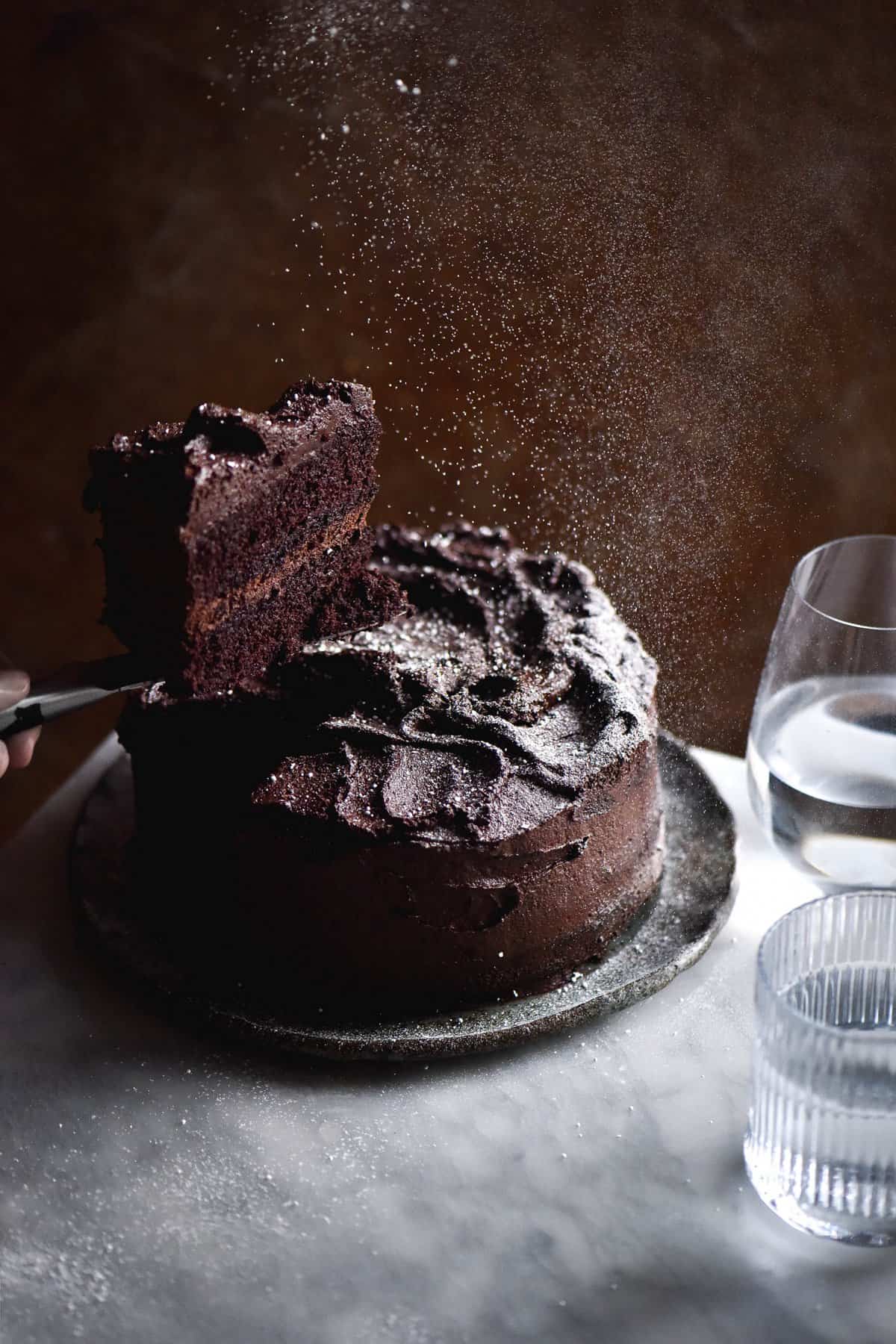 A gluten free chocolate cake sits atop a white marble table against a rusty brown backdrop. A slice of the cake has been removed and is held up by a hand from the left of the image. The whole cake is being sprinkled with snow-like icing sugar, and soft light filters through two water glasses to the right of the image