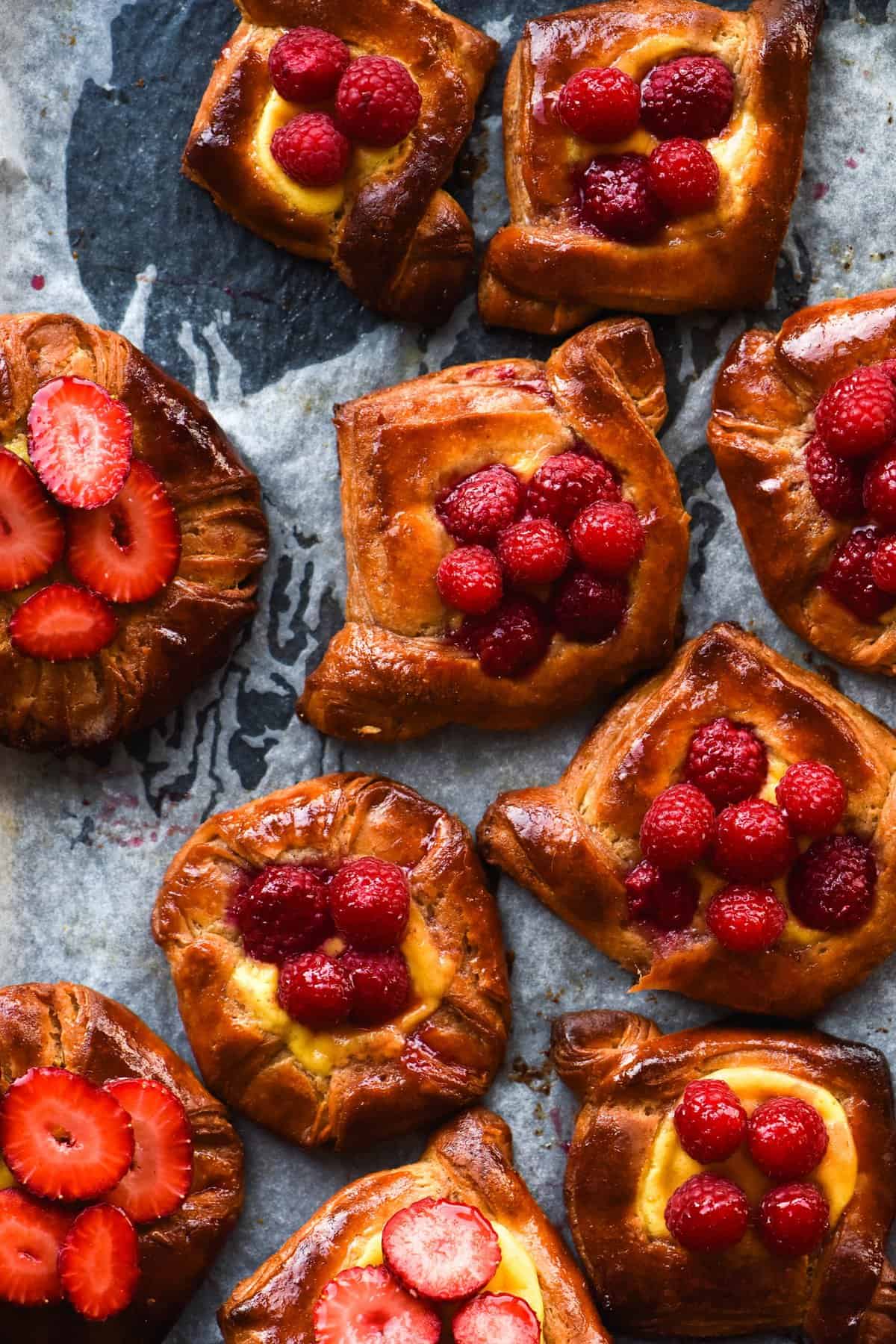 An aerial view of gluten free custard danishes topped with custard, vibrant strawberries and raspberries. The danishes sit atop a baking tray lined with baking paper