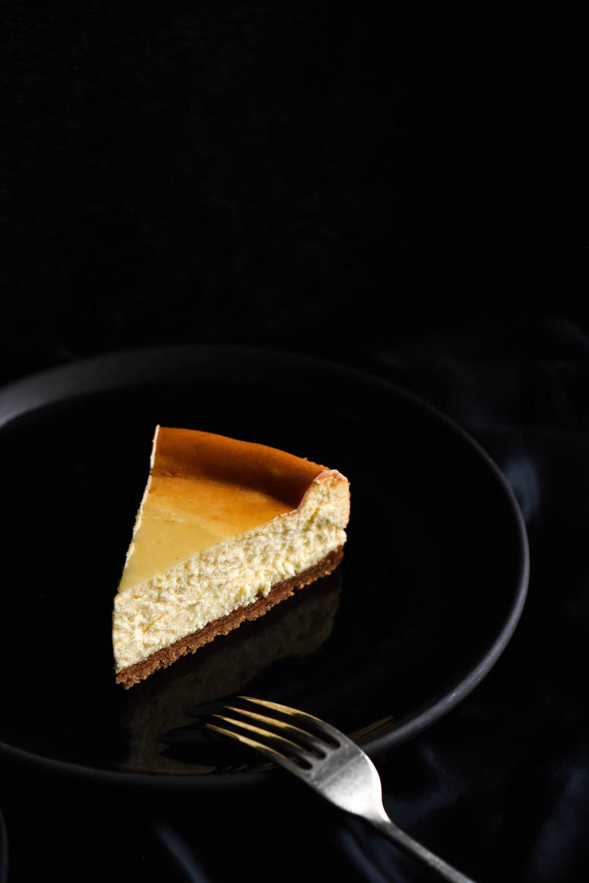 A dark and moody side on image of a slice of gluten free cheesecake on a black plate against a plate backdrop. A fork rests on the bottom right side of the plate. 