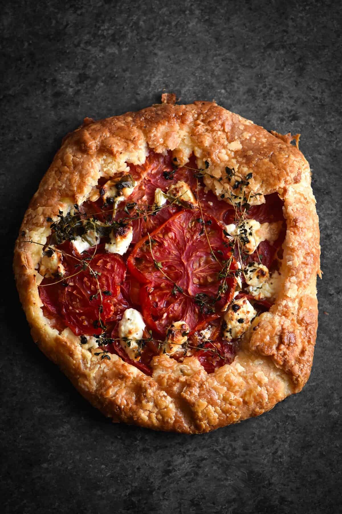 An aerial view of a gluten free tomato, thyme and goats cheese galette. The galette is golden brown and sits atop a dark grey metal backdrop