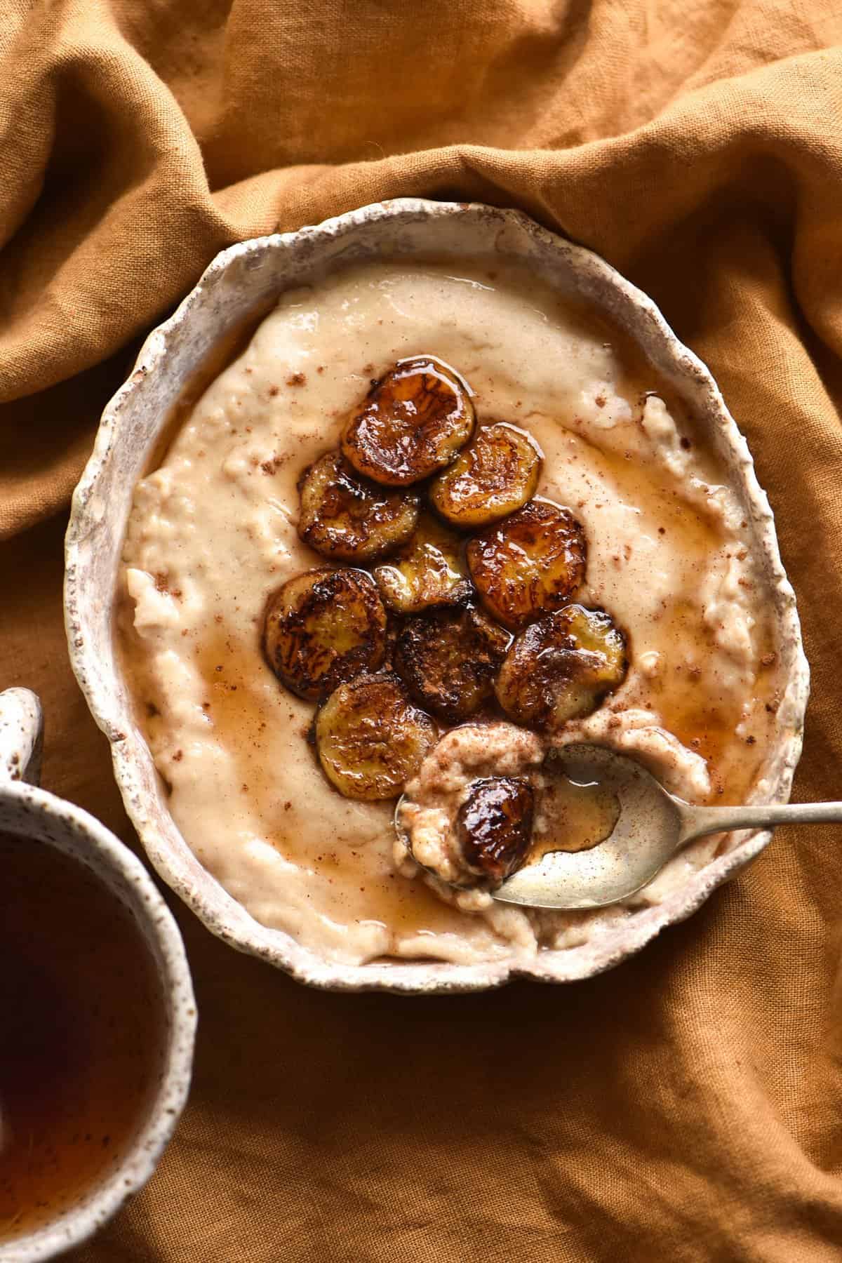 A bowl of gluten free, oat free porridge topped with caramelised bananas and cinnamon sits atop a rust coloured linen tea towel. A ceramic mug of tea sits to the bottom left of the image