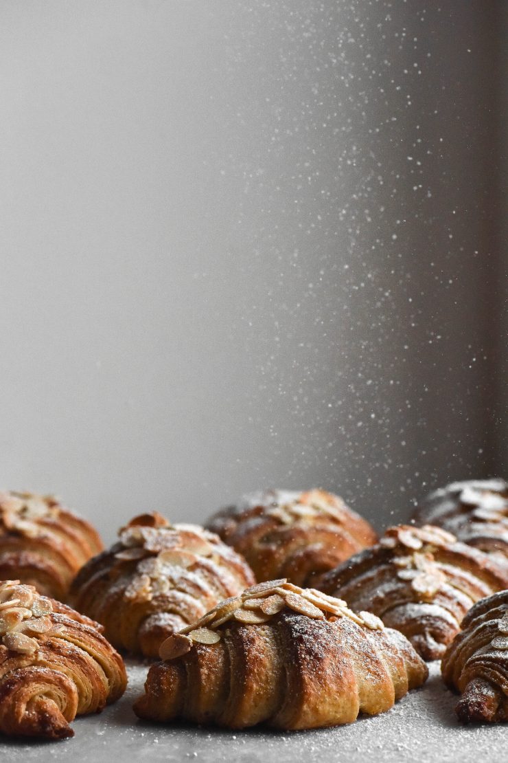 A side on view of gluten free almond croissants. The croissants are all facing the camera, exposing their lamination and beautiful layers. The croissants are being sprinkled with icing sugar that comes down from the top of the image