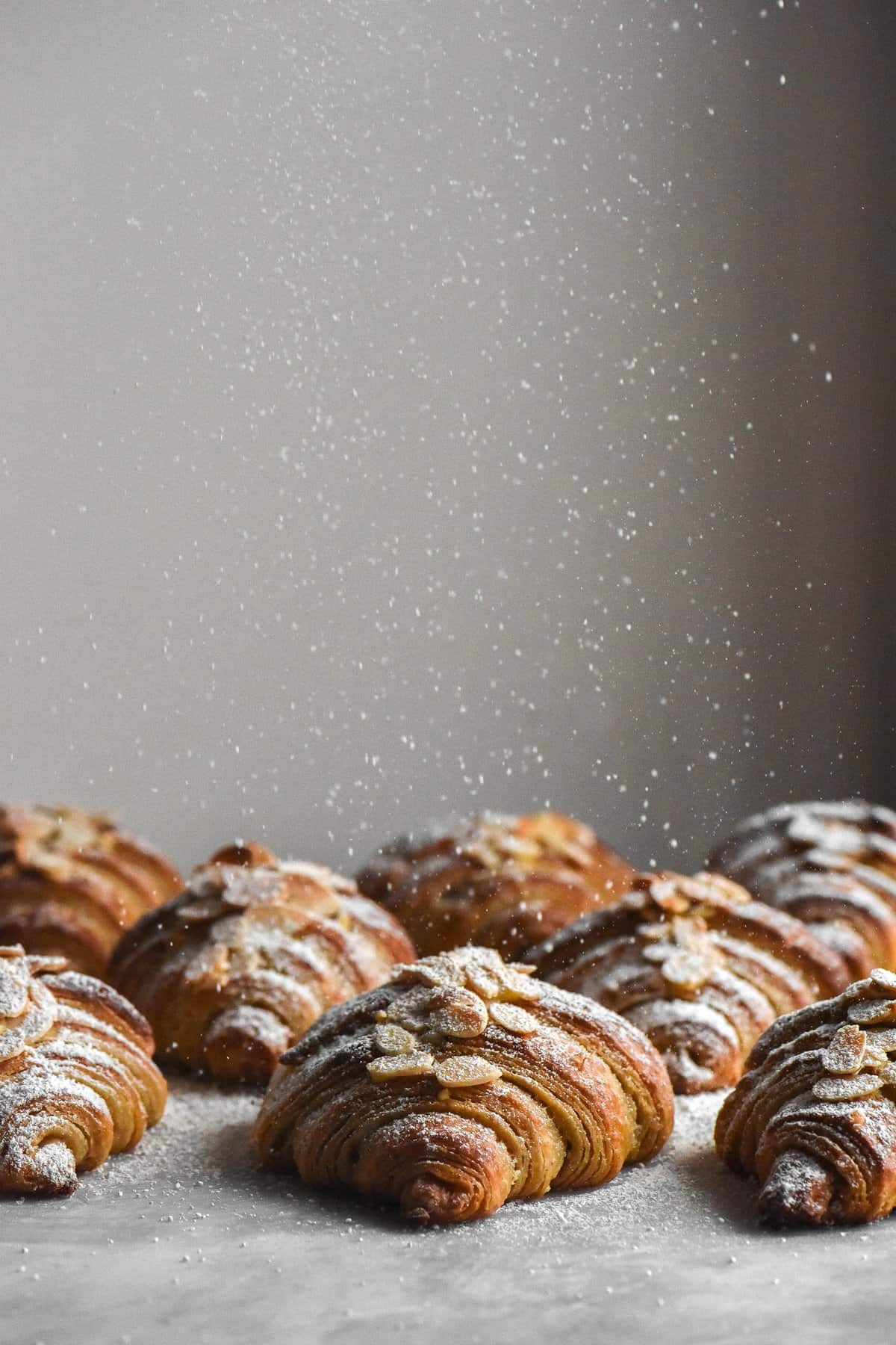 A side on view of gluten free almond croissants. The croissants are all facing the camera, exposing their lamination and beautiful layers. The croissants are being sprinkled with icing sugar that comes down from the top of the image