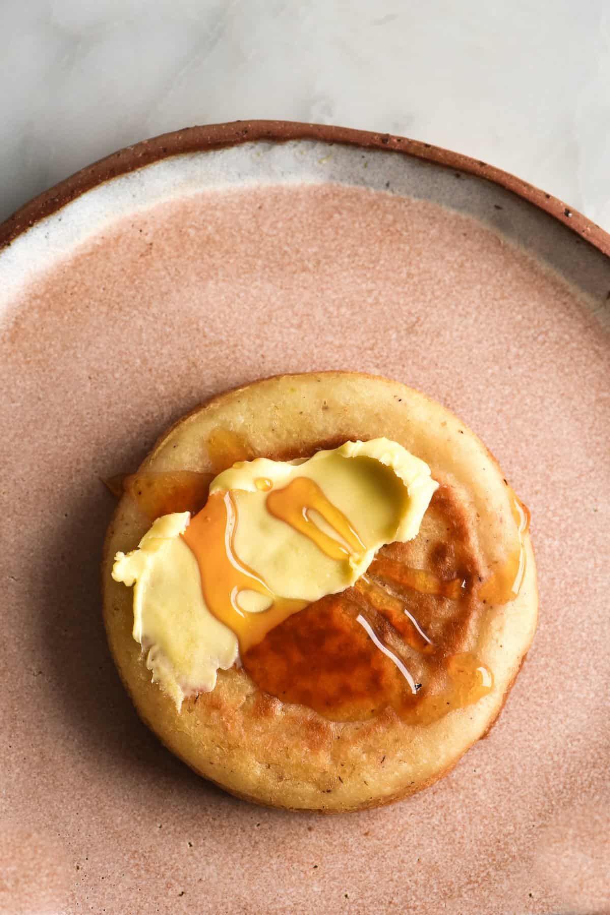 A gluten free crumpet sits atop a pale pink ceramic plate. The crumpet is topped with melting butter and a drizzle of honey