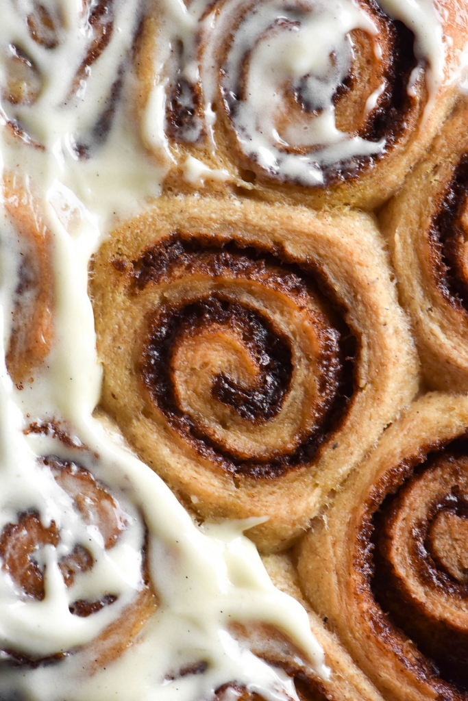 A close up aerial view of gluten free cinnamon scrolls, snug on a tray. The middle scroll is plain, but surrounded by scrolls smothered in low lactose cream cheese icing