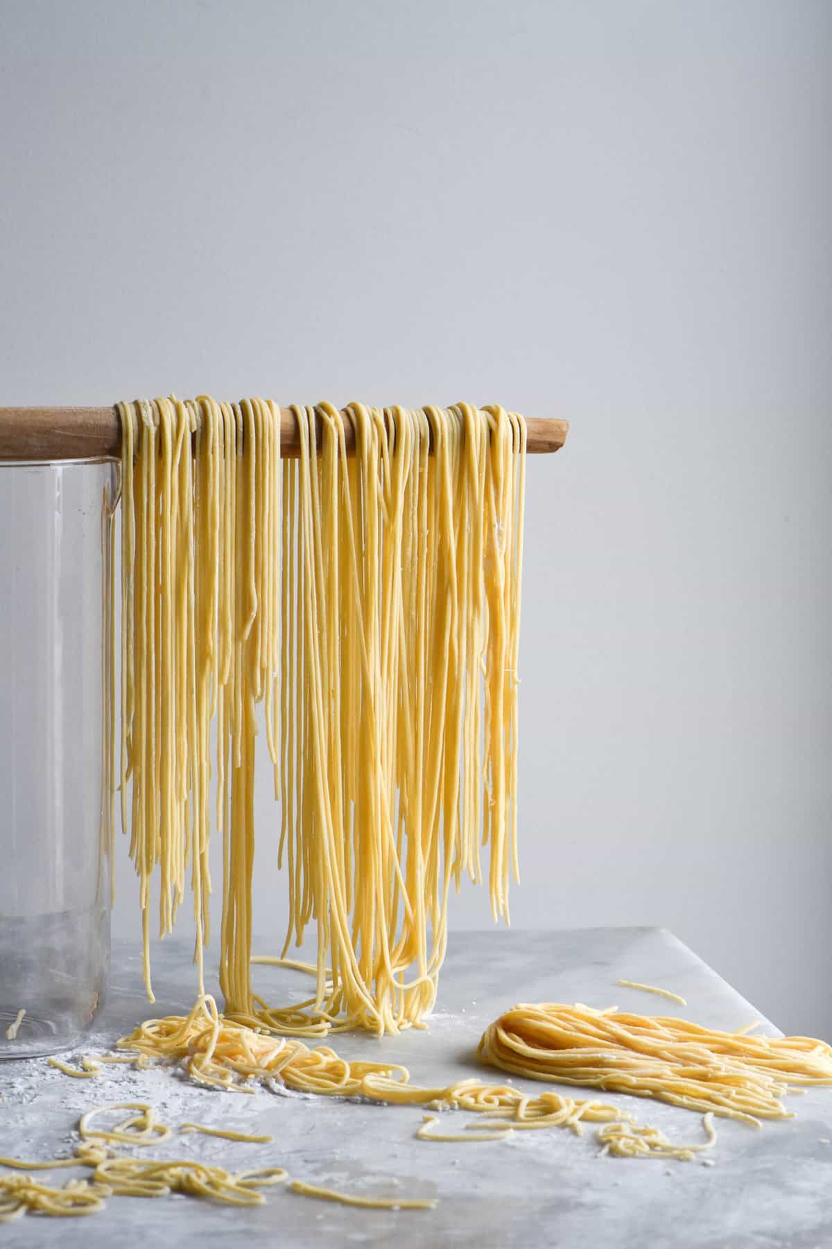 A side on view of gluten free egg noodles in the making. Set against a white marble table and white backdrop, noodles are draped over a spoon handle to dry as others are strewn across the table