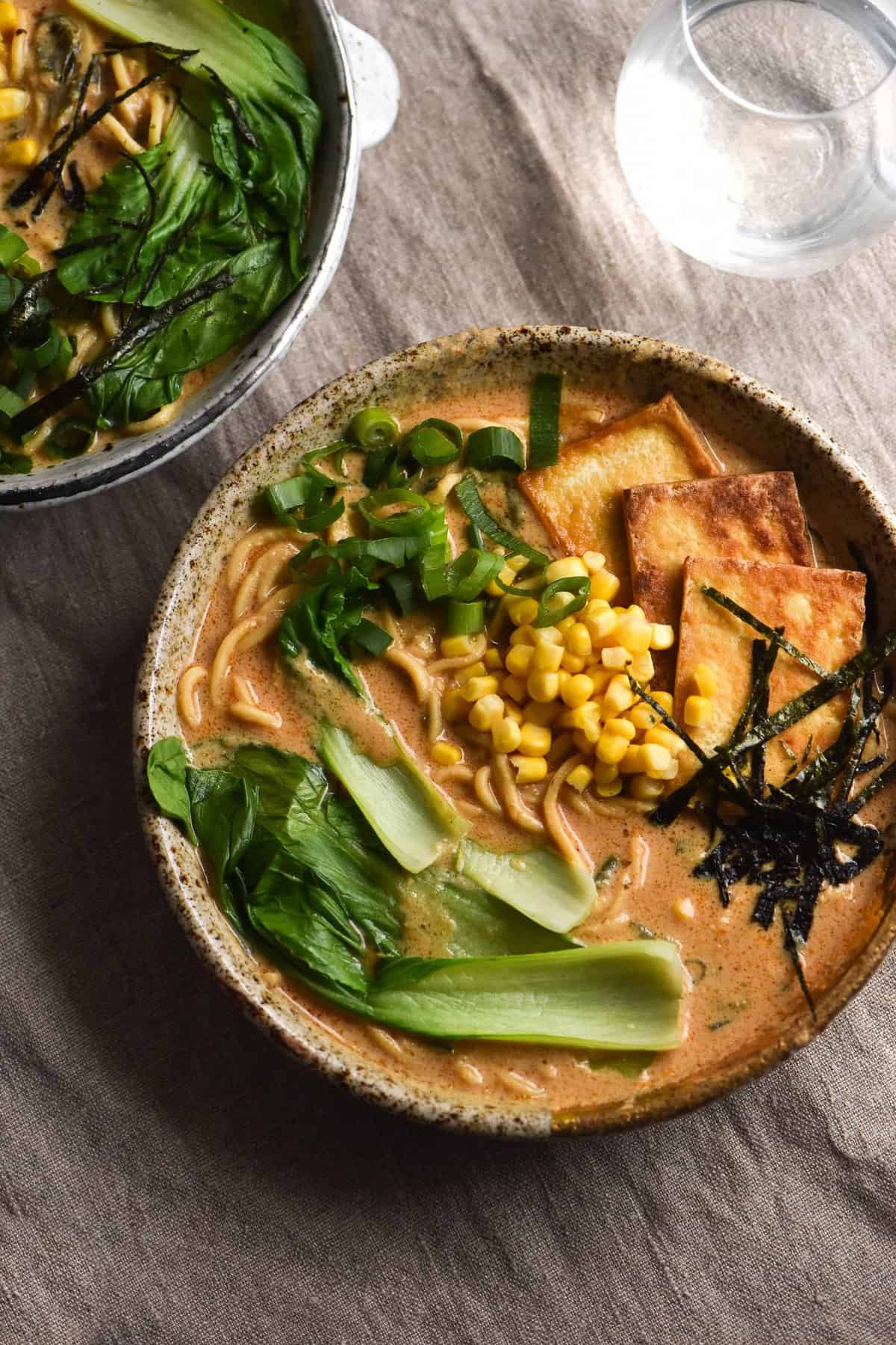 An aerial view of two bowls of FODMAP friendly, vegetarian or vegan and gluten free ramen. The bowls sit on a beige linen tea towel and a glass of water sits in the top right corner. The ramen is topped with smoked tofu slices, corn, spring onion greens, sliced nori and pak choi