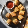 An aerial view of a plate of gluten free, vegetarian or vegan mini sausage rolls. The rolls sit atop a white ceramic plate on a white marble table, and a small bowl of tomato sauce sits in the top corner of the plate. The scene is lit up with harsh sunlight, creating light and shadow across the table