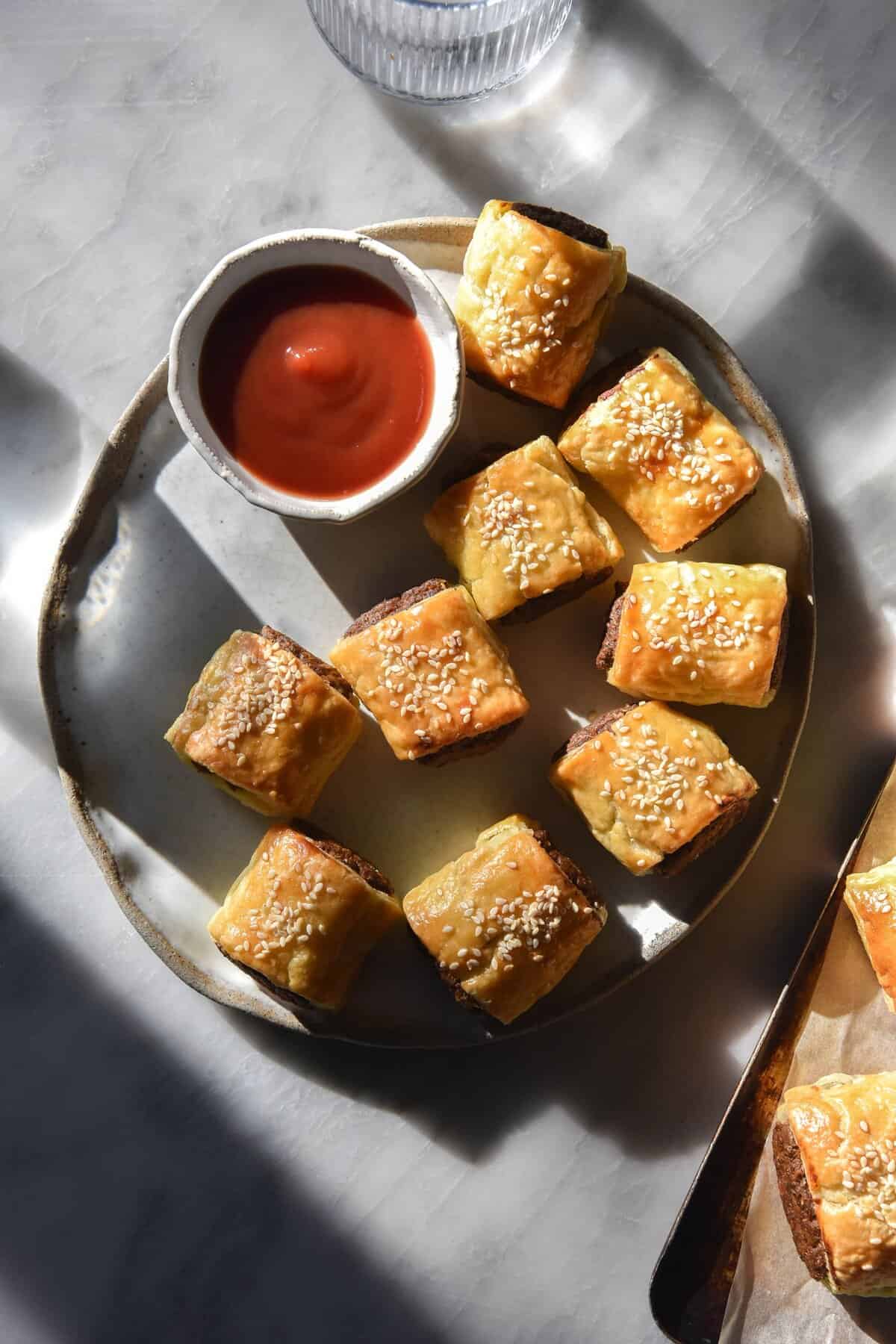 An aerial view of a plate of gluten free, vegetarian or vegan mini sausage rolls. The rolls sit atop a white ceramic plate on a white marble table, and a small bowl of tomato sauce sits in the top corner of the plate. The scene is lit up with harsh sunlight, creating light and shadow across the table