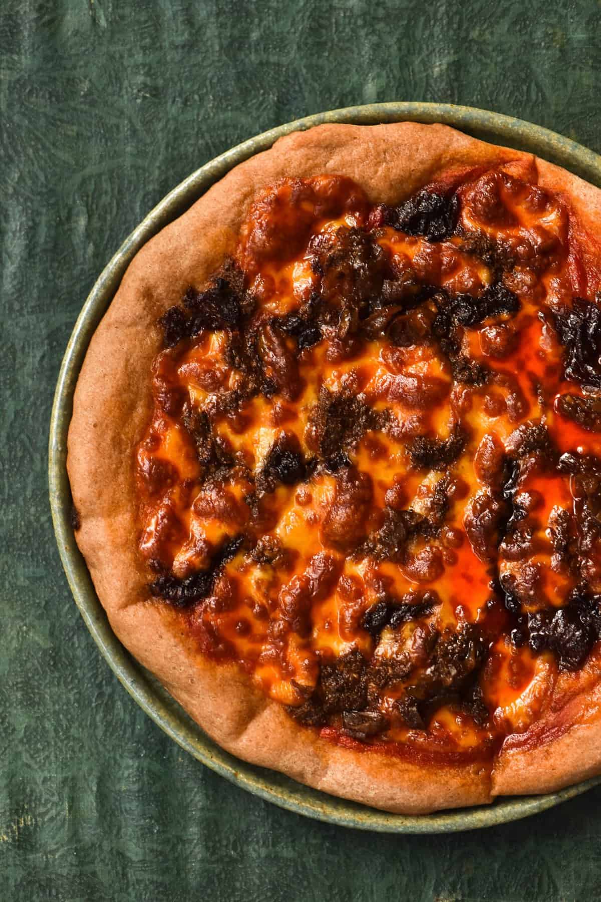 An aerial view of a gluten free, vegetarian sausage pizza with caramelised fennel and smoked mozzarella. The pizza sits atop a green ceramic plate against an olive green table top