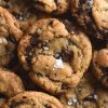 A close up aerial view of a stack of gluten free vegan choc chip cookies. The cookies are piled on top of each other, each topped with a sprinkle of sea salt flakes