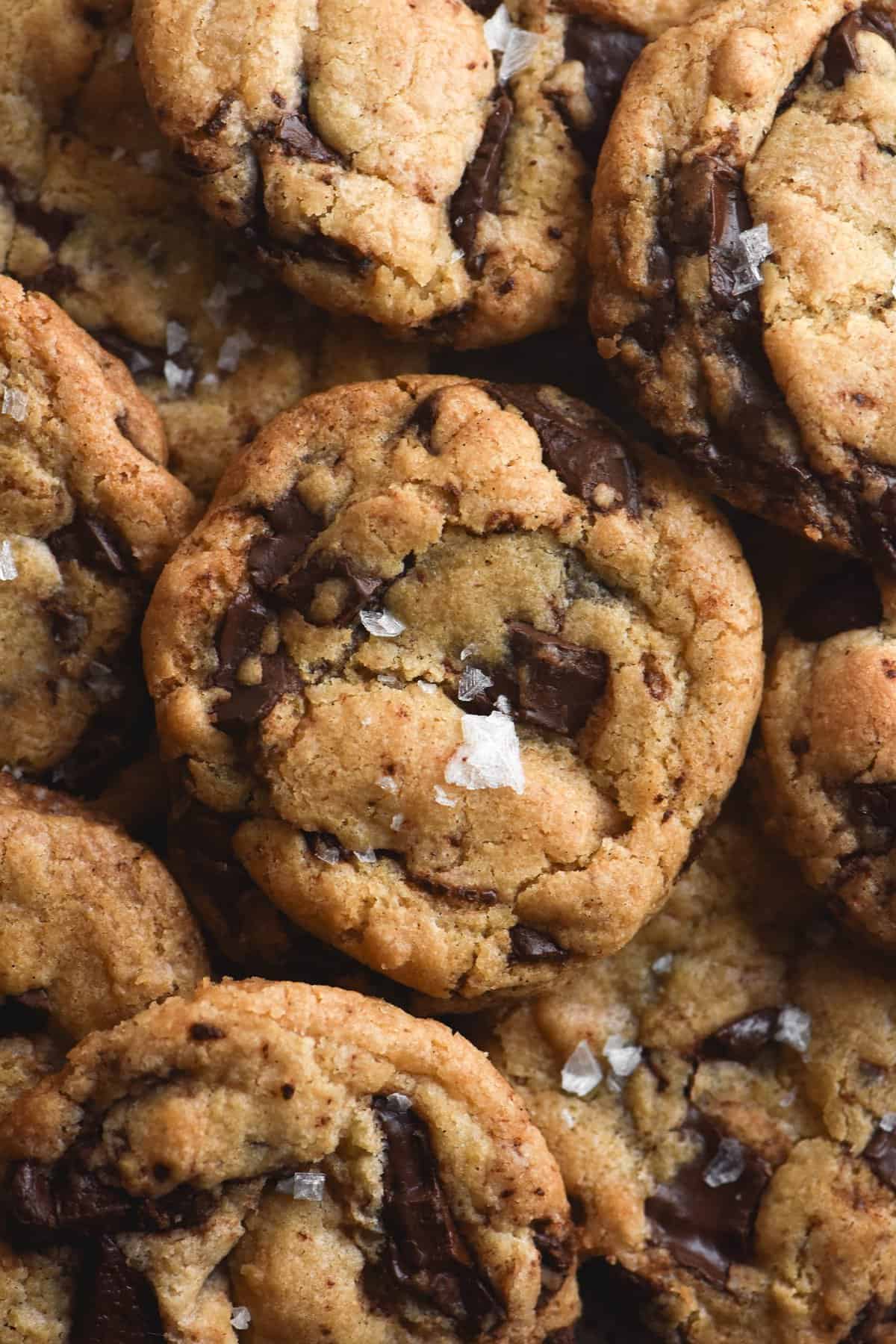 A close up aerial view of a stack of gluten free vegan choc chip cookies. The cookies are piled on top of each other, each topped with a sprinkle of sea salt flakes